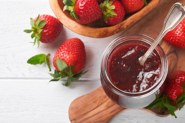 Confiture de fraises Confiture de fraises dans un bocal en verre avec assiette de baies fraîches sur fond de table en bois blanc gros plan Confiture fruitée aux fraises maison Vue de dessus avec espace de copie