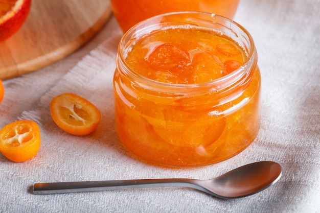 Confiture d’orange et de kumquat dans un bocal en verre avec des fruits frais sur une nappe en lin blanc.