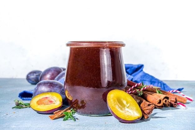 Confiture de prunes d'automne épicée dans un petit pot, avec de la cannelle, du thym, de l'anis et des prunes fraîches sur fond bleu béton