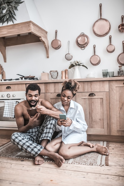 Confort de la maison. Femme focalisée à la peau sombre dans les vêtements à la maison pieds nus avec smartphone et mari mangeant assis sur le sol de la cuisine