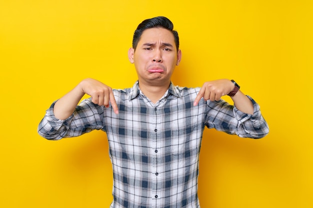 Confus jeune bel homme asiatique portant une chemise à carreaux pointant les doigts vers le bas montrant un endroit libre pour la publicité isolé sur fond jaune Concept de style de vie des gens