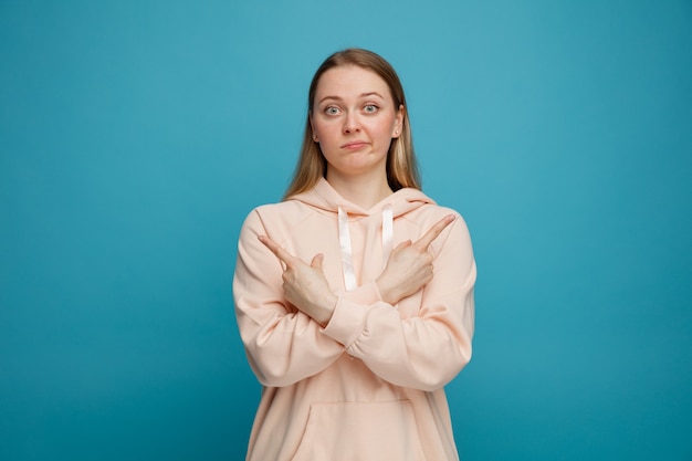 Confus jeune femme blonde en gardant les mains croisées pointant vers les côtés