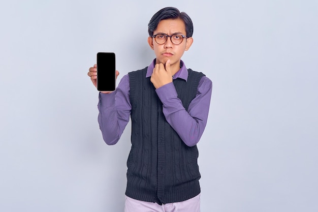 Confus jeune homme asiatique en chemise décontractée et gilet montrant un écran vide de téléphone portable recommandant l'application isolée sur fond blanc Concept de style de vie des gens