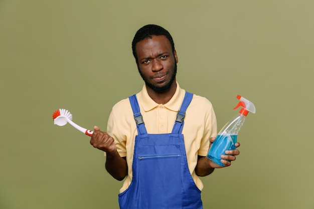 Confus tenant un agent de nettoyage avec une brosse de nettoyage jeune homme nettoyant afro-américain en uniforme avec des gants isolés sur fond vert