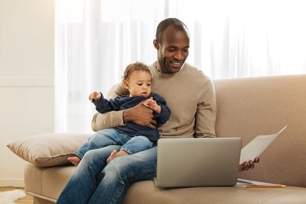 Congé paternel. Enthousiaste afro-américain barbu aux yeux sombres souriant et tenant son fils sur ses genoux tout en travaillant sur l'ordinateur portable et tenant une feuille de papier