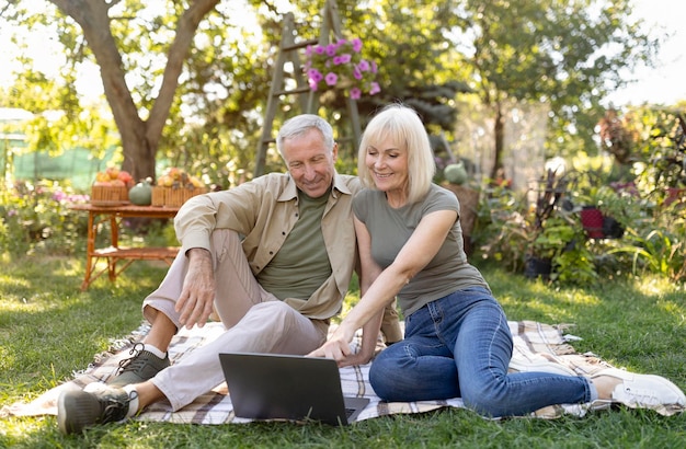 Conjoints seniors modernes faisant leurs achats en ligne avec un ordinateur portable assis sur une couverture dans le jardin profitant d'un printemps chaud