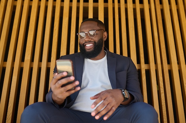 Connecté. Un homme à la peau foncée en costume assis sur le banc avec un smartphone dans les mains