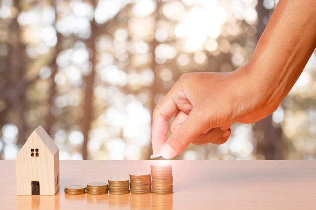 Économiser de l'argent à la main en mettant des pièces sur une pile avec un modèle de maison en bois sur fond de feuille floue.