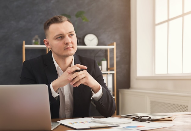 Conseiller financier masculin sérieux travaillant avec des papiers et un ordinateur portable à l'intérieur d'un bureau moderne. Homme pensif en tenue de soirée vérifiant les documents et réfléchissant à la stratégie, espace de copie