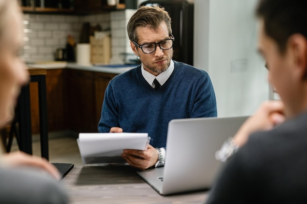 Conseiller financier sérieux écrivant des notes et utilisant un ordinateur portable lors d'une réunion avec un couple à leur domicile