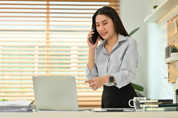 Conseillère en placement asiatique ayant une conversation téléphonique consultant un client et utilisant un ordinateur portable sur le lieu de travail