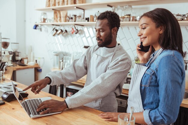 Conseils nécessaires. Charmant barista masculin debout à côté de sa collègue derrière le comptoir et lui demandant des conseils sur la mise à jour du menu sur leur site Web pendant que la femme appelle quelqu'un