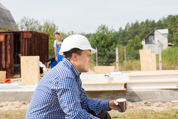 Constructeur assoiffé prenant une pause-café assis vue latérale avec une tasse de café dans les mains pendant que son équipe continue de travailler sur le bâtiment derrière lui