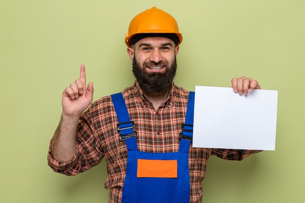 Constructeur barbu en uniforme de construction et casque de sécurité tenant une page blanche à la recherche de sourire gaiement montrant l'index ayant une nouvelle idée