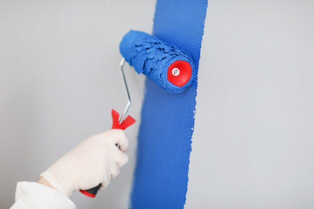 Constructeur dans des gants de protection peignant le mur blanc bleu avec le plan rapproché de rouleau