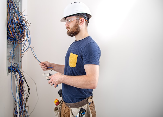 Constructeur électricien au travail, examine la connexion du câble dans la ligne électrique dans le fuselage d'un tableau industriel. Professionnel en salopette avec un outil d'électricien.