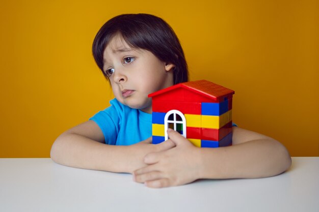 Constructeur d'enfant garçon dans un T-shirt bleu contre un mur jaune construit une maison à partir de cubes en plastique colorés.