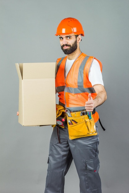 Un constructeur d'hommes dans un casque orange avec une boîte en carton dans ses mains Sur un fond gris