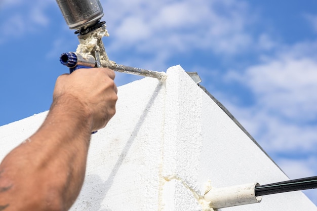 Le constructeur isole les murs de la maison avec de la mousse de polystyrène et comble les lacunes avec de la mousse de polyuréthane Un pistolet avec de la mousse dans les mains d'un constructeur contre un ciel bleu