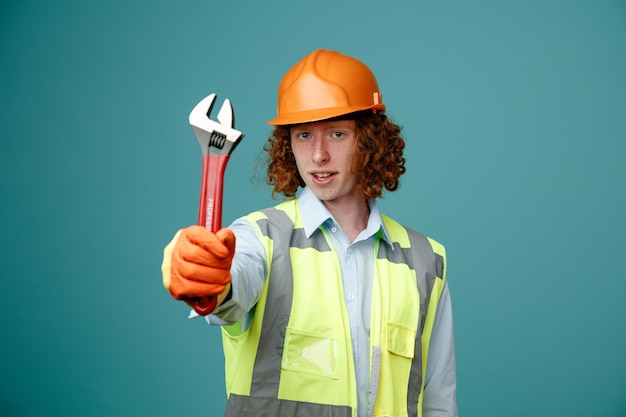 Constructeur jeune homme en uniforme de construction et casque de sécurité montrant une clé regardant la caméra avec un visage sérieux debout sur fond bleu