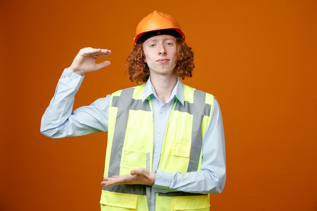 Constructeur jeune homme en uniforme de construction et casque de sécurité regardant la caméra avec un sourire confiant montrant le symbole de taille avec les mains debout sur fond orange