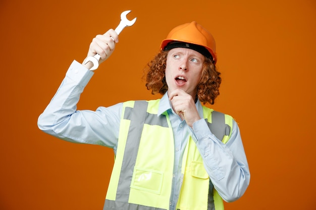 Constructeur jeune homme en uniforme de construction et casque de sécurité tenant une clé en levant avec une expression pensive pensant debout sur fond orange