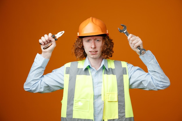 Constructeur jeune homme en uniforme de construction et casque de sécurité tenant une clé et une pince regardant la caméra avec un visage en colère ennuyé debout sur fond orange