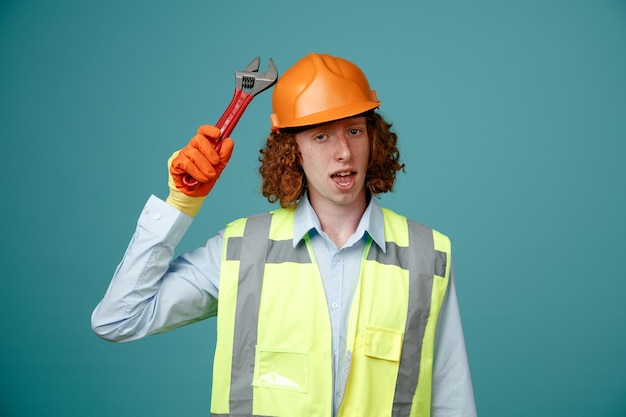 Constructeur jeune homme en uniforme de construction et casque de sécurité tenant une clé regardant la caméra confus debout sur fond bleu