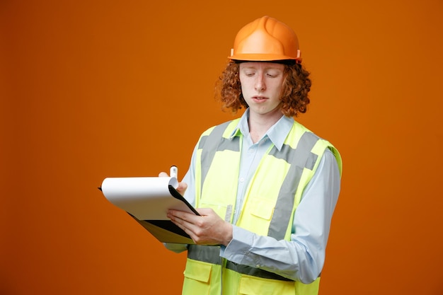 Constructeur jeune homme en uniforme de construction et casque de sécurité tenant le presse-papiers et le marqueur écrivant quelque chose qui a l'air confiant debout sur fond orange