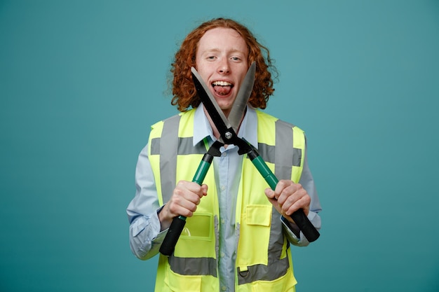 Photo constructeur jeune homme en uniforme de construction tenant des ciseaux s'amusant à couper sa langue debout sur fond bleu