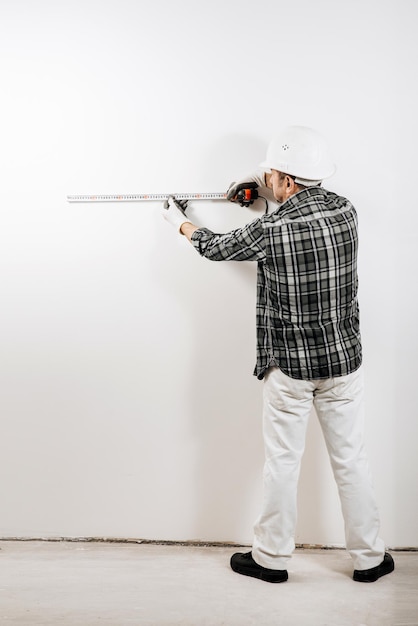 Un constructeur masculin dans un casque mesure un mur blanc avec un ruban à mesurer de construction