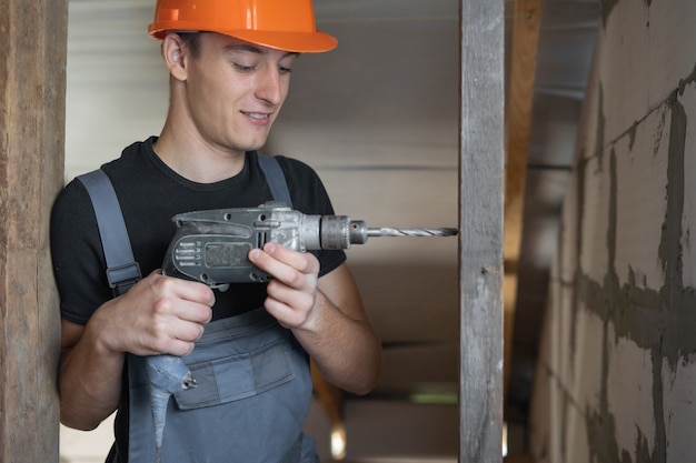 Constructeur masculin vêtu de vêtements de travail et d'un casque orange. Tient une perceuse et fait un trou dans un morceau de bois. Espace de copie