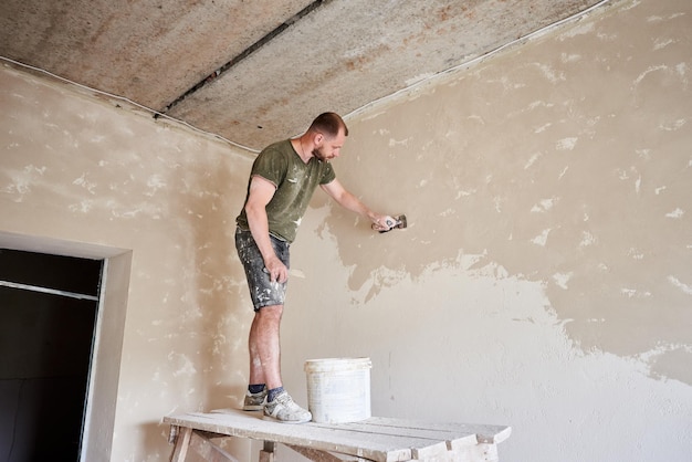 Le constructeur se tient sur un support en bois et travaille avec une spatule avec du plâtre au mur dans une petite pièce