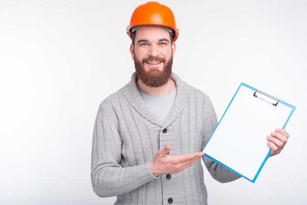 Un constructeur souriant et pointant sur un porte-papier près de fond blanc.