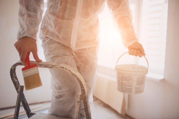 Le Constructeur Travaille Sur Le Chantier Et Mesure Le Plafond. Travailleur Avec Seau Et Rouleau à Peinture Près Du Mur.