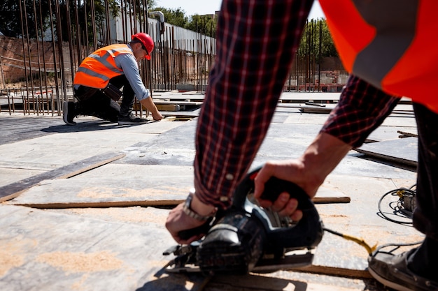 Le constructeur vêtu d'un gilet de travail orange et d'un casque utilise une scie circulaire sur le chantier ouvert. .