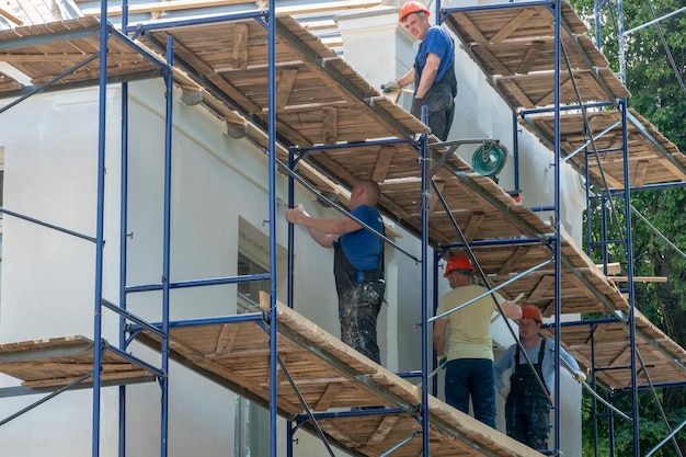 Les constructeurs debout sur des échafaudages sont engagés dans la réparation de la façade d'un bâtiment Plâtre et peinture de la façade de la maison L'équipe de construction travaille dur dans de mauvaises conditions de travail