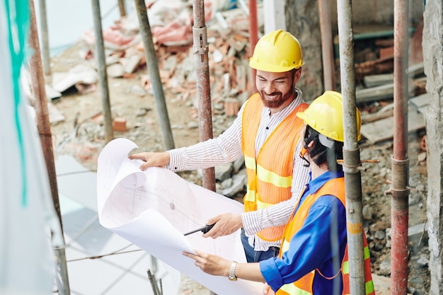 Constructeurs positifs en casque jaune vérifiant le plan du bâtiment et discutant du plan ou des workrs