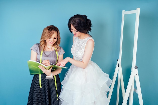La consultante en mariage couturière et la mariée discutent des détails de la robe de mariée en studio sur fond bleu. couturière sur mesure et bal des finissants dans le dressing à côté du miroir.