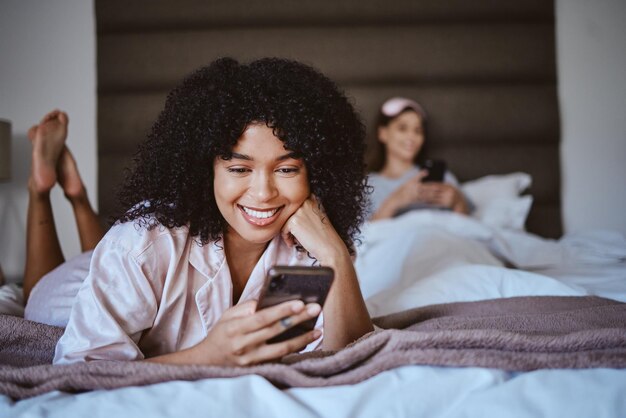 Contacter le téléphone et sourire avec une femme noire lors de la soirée pyjama pour la communication sur Internet ou les rencontres en ligne Bonne détente et textos avec une fille et des amis dans la chambre avec la technologie des médias numériques ou sociaux