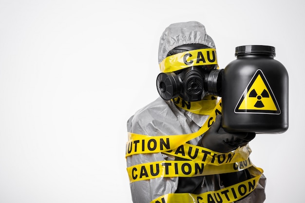 Contamination radioactive. Un chimiste scientifique vêtu d'une tenue de protection, enveloppé dans un ruban jaune "attention", tient dans ses mains un récipient noir avec un signe de danger de radiation. Espace copie