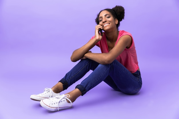 Contenu attrayant femme afro-américaine souriant et parlant sur smartphone, assis sur le sol, isolé