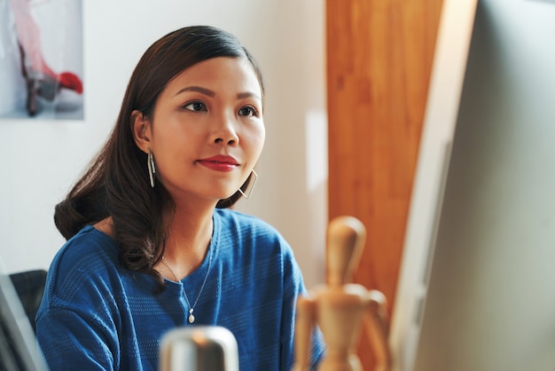 Contenu belle jeune créatrice asiatique en boucles d'oreilles assise dans un bureau contemporain et travaillant avec un ordinateur