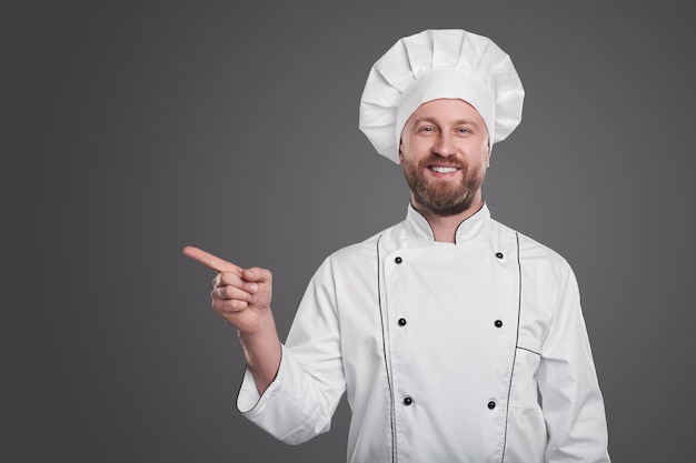 Contenu cuisinier homme barbu en uniforme blanc avec tuque montrant loin tout en regardant la caméra