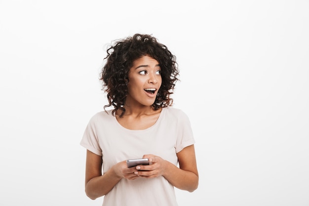 Contenu femme brune avec une coiffure afro à l'aide de smartphone et à côté sur fond, isolé sur mur blanc