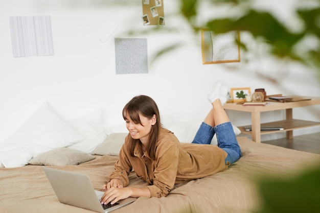 Contenu fille indépendante aux cheveux bruns allongée sur le lit et tapant sur un ordinateur portable tout en travaillant à la maison