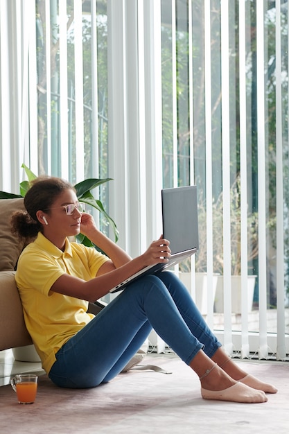 Contenu fille de race mixte dans des écouteurs sans fil et des lunettes assis sur le sol avec une tasse de jus et utilisant un ordinateur portable tout en regardant le webinaire
