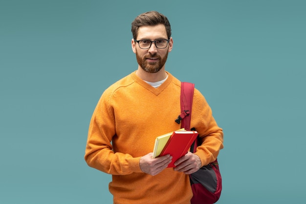 Contenu jeune étudiant homme en tenue décontractée regardant la caméra tout en posant avec sac à dos et manuel isolé sur fond bleu