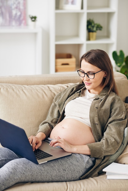 Contenu jeune femme enceinte à lunettes assis sur un canapé confortable et travaillant avec un ordinateur portable en utilisant le wi-fi à la maison