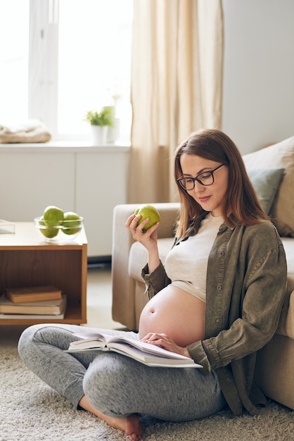 Contenu jeune femme enceinte à lunettes assis avec les jambes croisées sur le tapis et manger une pomme verte en lisant un livre à la maison